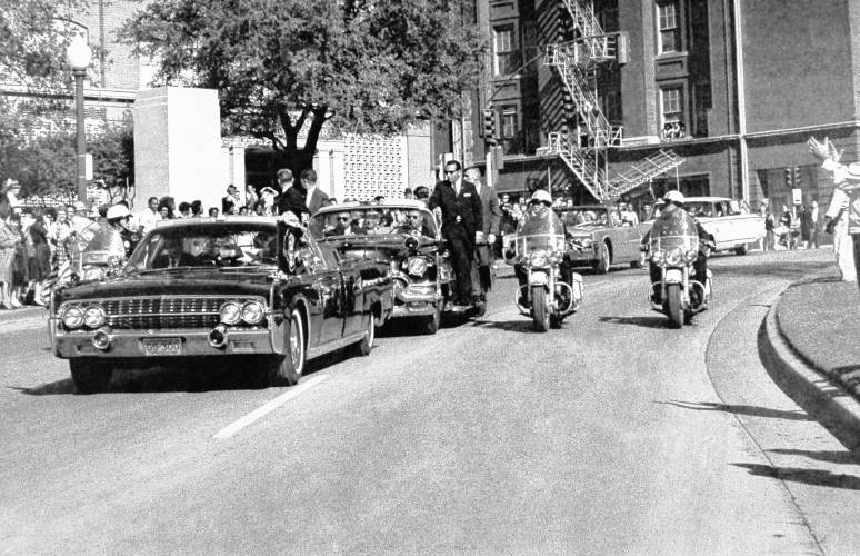 This photograph captures the moment the first shot hits the president. Seen through the limousine’s windshield, the president appears to raise his hand toward his head. Mrs. Kennedy’s gloved left hand reaches out to hold him. The two secret service agents riding the right running board of the second car turn in the direction of the gunfire.