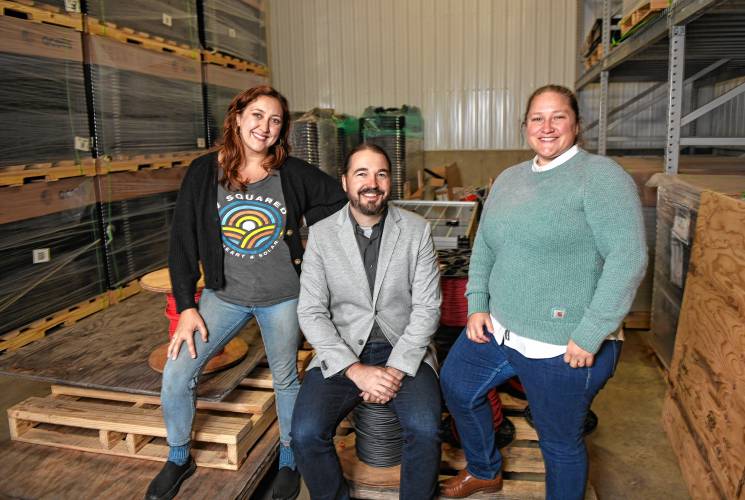 Workers-owners Brittany Hathaway, Josh Hilsdon and Kate Carter of PV Squared with stacks of solar panels and spools of wire in the company’s Greenfield warehouse.