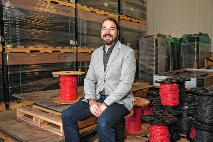 Josh Hilsdon of PV Squared with stacks of solar panels and spools of wire in the company’s Greenfield warehouse.